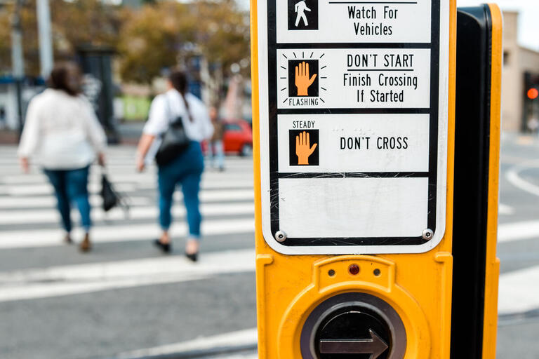 a crosswalk pedestrian signal button in the foreground; two people crossing the street in the background